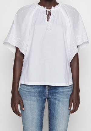 Osoba oblečená v bílém blouse s vyšívanými širokými rukávy a zavazovacími krčními šňůrkami, kombinované s modrými vybledlými džíny.