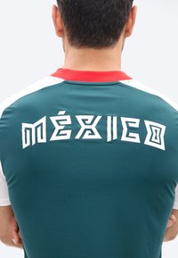 Muž v zeleném sportovním dresu s červeným límcem a bílými rukávy, na zádech je stylizovaný nápis „MÉXICO“ bílými písmeny.