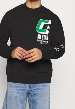 Osoba nosząca czarną bluzę Converse All Star z zielono-białym logo na piersi i białymi wzorami gwiazd na lewym rękawie.