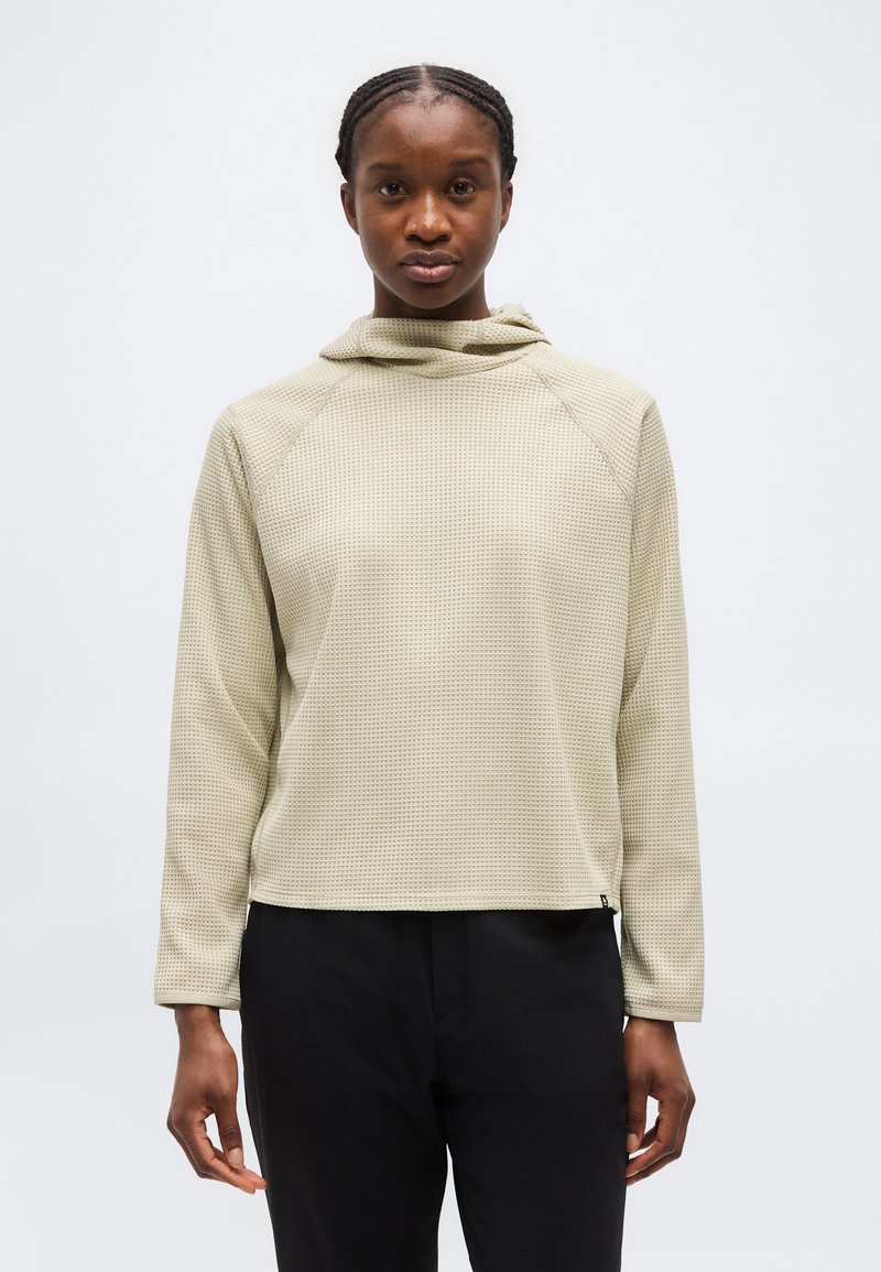 Άτομο με πλεξούδες, φορώντας μπεζ υφασμένη μακρυμάνικη μπλούζα με κουκούλα και μαύρο παντελόνι, που κοιτάει μπροστά μπροστά σε ένα απλό ανοιχτό φόντο.