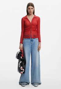 Rotes, transparentes, langärmliges Zip-Top mit einem strukturierten Muster, kombiniert mit weit geschnittenen, hellblauen Jeans und einer schwarzen Handtasche mit Blumenmuster.