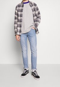 Hombre con camiseta gris claro, camisa a cuadros en blanco y negro, jeans azul claro y zapatillas negras, de pie frente a un fondo blanco liso.