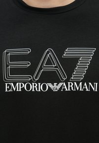 Mustad lühikeste varrukatega t-särk, millel on valged tekstid: "EA7" ja "EMPORIO ARMANI" stiilsetes fontides ning tiibadele sarnanev logo. Sile kangapind.