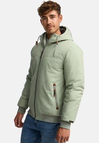 Man wearing light green hooded puffer jacket with zipper and zippered side pocket, paired with blue jeans, standing against white background.