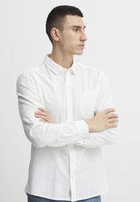 Camisa blanca de manga larga y botones, hecha de una tela ligera y texturizada. Presenta un bolsillo en el pecho y puños abotonados.