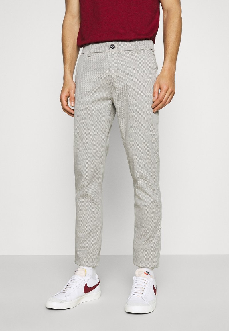 Homme portant un pantalon slim gris clair, des baskets blanches avec un swoosh rouge de Nike, et une chemise bordeaux, se tenant devant un fond uni.