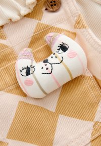 Jouet en peluche en forme de croissant avec des traits du visage brodés, présentant des couleurs pastel douces, reposant sur un fond en tissu à motifs.