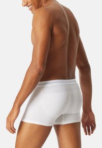 Mandlig model med medium hudfarve iført hvide boxer shorts, set bagfra med et let smil, mod en ensfarvet baggrund.