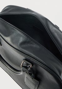 Sac en cuir noir avec une texture lisse, ouverture zippée et détail de poignée torsadée. L'intérieur est sombre avec une étiquette simple.