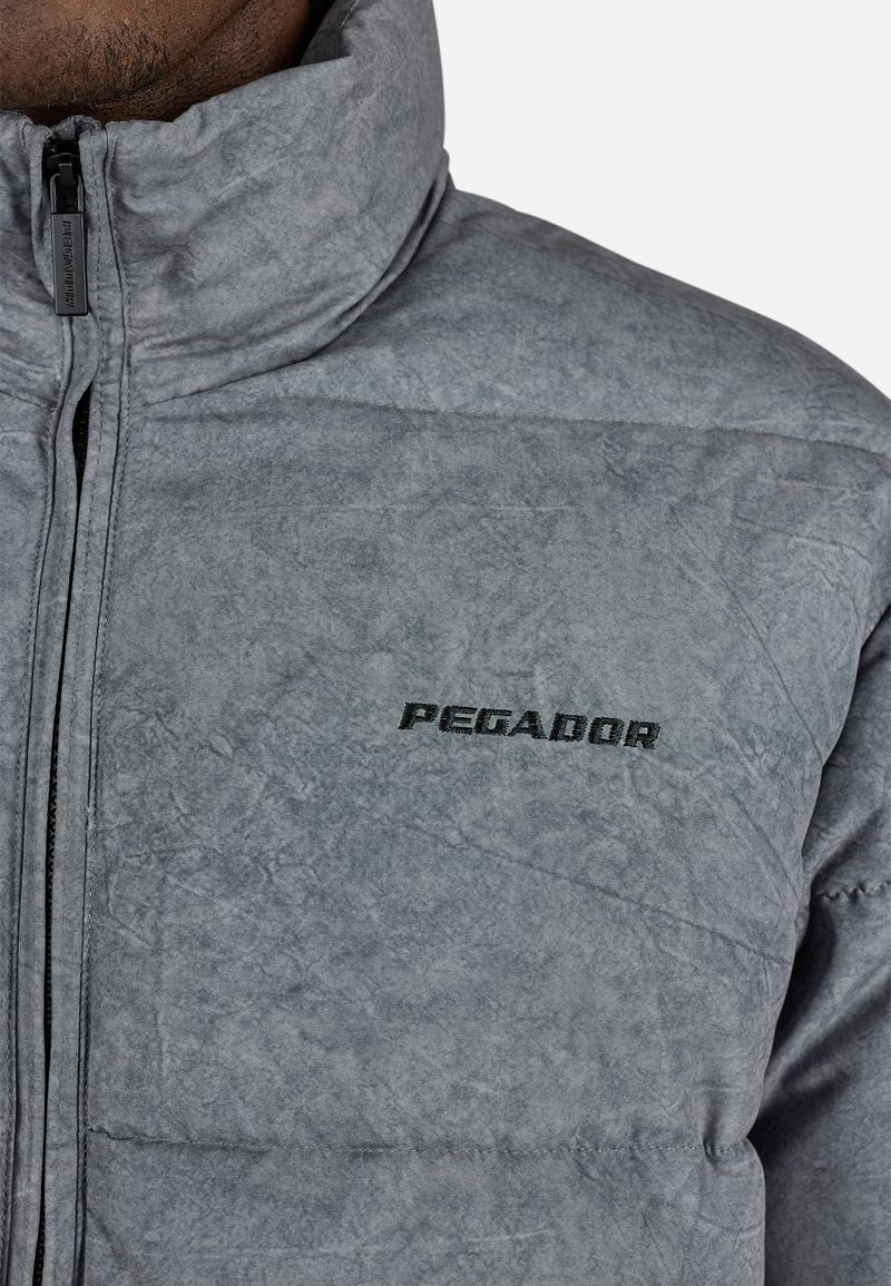 Chaqueta acolchada gris con cuello alto, que presenta una textura suave y un ligero brillo. Logo negro "PEGADOR" en el pecho. Cierre de cremallera frontal completo.