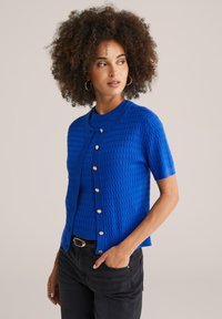 Cardigan en tricot bleu à manches courtes avec un motif texturé, encolure ronde et boutons dorés. Porté par-dessus un jean foncé.