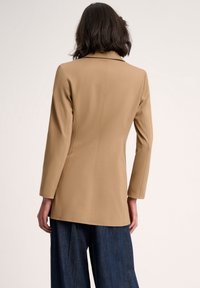 Hellbrauner taillierter Blazer mit Reverskragen, langen Ärmeln und einer strukturierten Passform. Glatter Stoff mit dezenten Nahtdetails und leichter Textur.