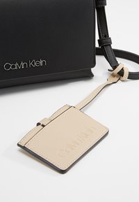 Černá kabelka Calvin Klein s přišitou béžovou koženou visačkou, na které jsou vyražené a metalické loga značky.