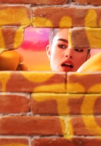 Joven con labios pintados de rojo asoma a través de un agujero irregular en una pared de ladrillo cubierta de graffiti hacia un cielo de atardecer colorido.