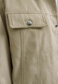 Beige Kunstlederjacke mit einer knöpfbaren Brusttasche, subtilen Nähten und einer glatten Textur.
