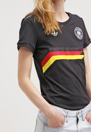 Personne portant un t-shirt Adidas noir avec des rayures chevrons rouges, jaunes et noires et un emblème, accompagné d'un jean bleu clair, mains dans les poches.