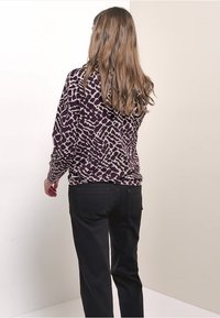 Eine Frau mit langen hellbraunen Haaren trägt ein langärmliges Oberteil mit abstraktem Muster in Lila und Beige sowie schwarze Hosen und steht vor einer schlichten Wand.