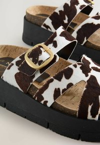 Sandalias de plataforma de estampado de vaca marrón y blanco con suela de corcho, base de goma negra y un acento de hebilla dorada en la correa.