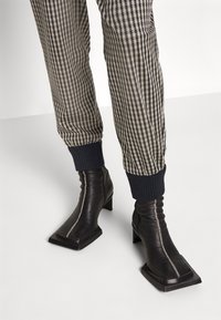 Bottes noires en cuir à la cheville avec un bout carré et un talon bloc, arborant un détail de couture visible. Assorties avec un pantalon à motif écossais.
