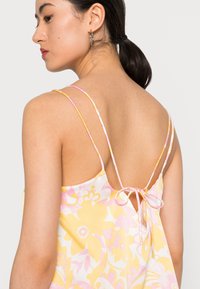 Vestido floral com cores amarelo, rosa e branco, com alças finas e detalhe nas costas com laço. Tecido suave com um leve brilho.
