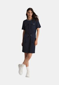 Abito blu navy con maniche corte, due tasche frontali con bottone, vita con cordoncino e vestibilità comoda, abbinato a scarpe da ginnastica bianche e calze.