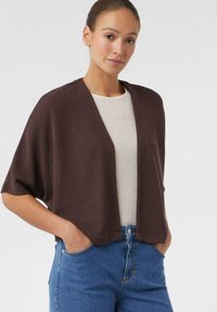 Bruine gebreide cardigan met korte mouwen, open voorkant en een relaxte pasvorm, gecombineerd met een licht textuurtop en blauwe denim jeans.