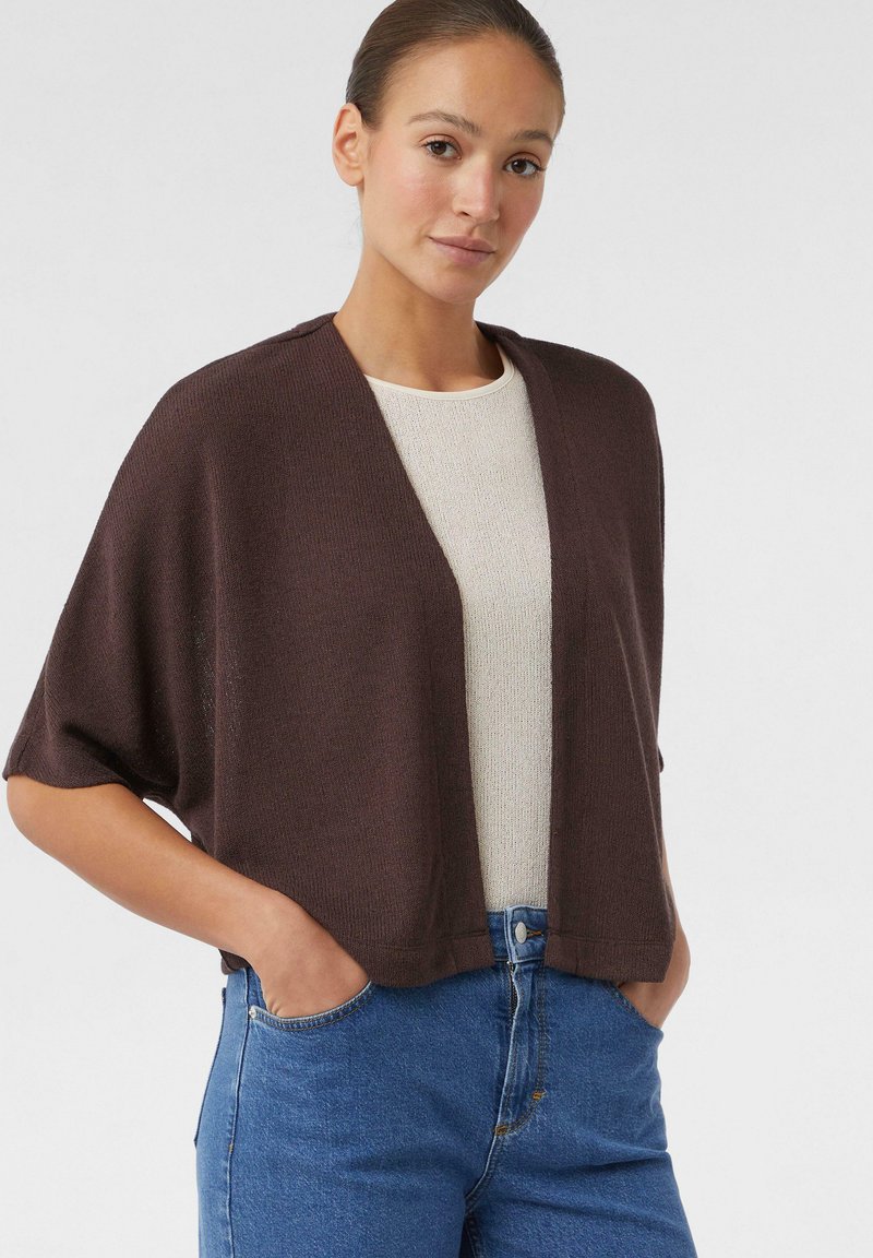 Bruine gebreide cardigan met korte mouwen, open voorkant en een relaxte pasvorm, gecombineerd met een licht textuurtop en blauwe denim jeans.