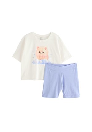 Teenagepige stående barfodet iført en hvid cropped t-shirt med et katte-design og lyseblå cykelshorts mod en hvid baggrund.