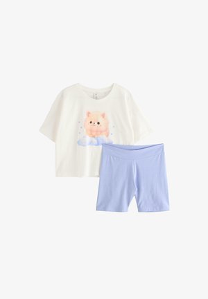 Tienermeisje staat op blote voeten, draagt een wit cropped T-shirt met een kattenmotief en lichtblauwe biker shorts tegen een witte achtergrond.