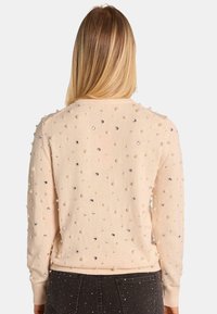 Beige Pullover mit runden Perlen- und Schmuckverzierungen, gerippten Bündchen und Saum sowie Rundhalsausschnitt. Die Textur wirkt weich und gestrickt.
