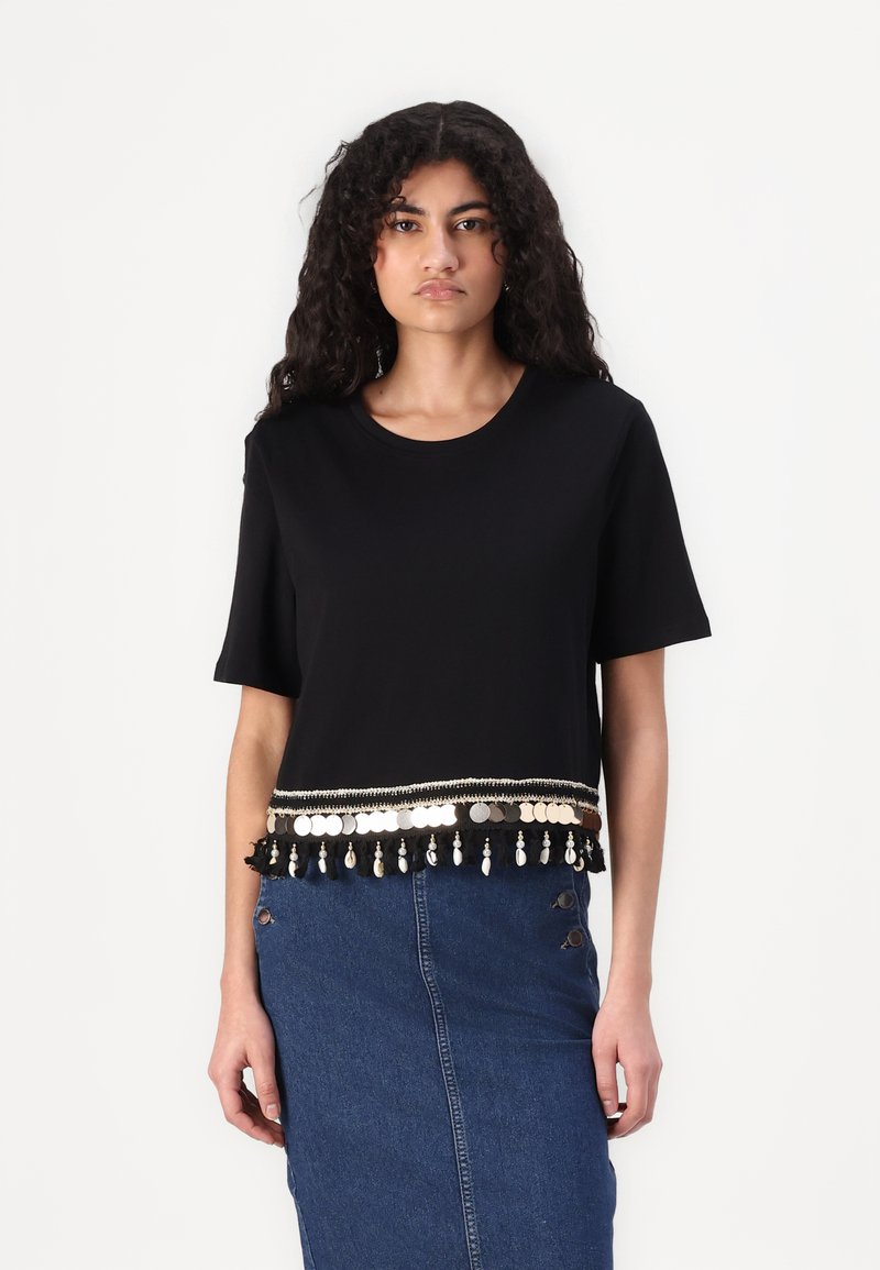 Camiseta negra corta de algodón, con un borde decorativo con acentos metálicos y flecos en el dobladillo, combinada con una falda de mezclilla.