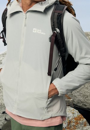 Person trägt eine hellgraue Jack Wolfskin Jacke mit Frontreißverschluss und schwarze Rucksackgurte, steht draußen nahe Felsen.