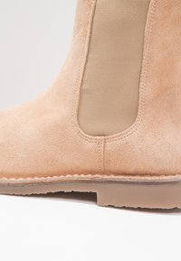 Botín de ante beige a la altura del tobillo con un panel lateral elástico liso y una suela de color marrón cosida. Presenta una punta redonda y un acabado texturizado.