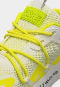 Λευκό και κίτρινο αθλητικό παπούτσι από διχτυωτό ύφασμα με κίτρινες κορδέλες, λογότυπο "Versace Jeans Couture" στη γλώσσα και στο πλευρό της σόλας.