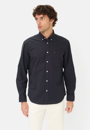 Mężczyzna ubrany w dopasowaną czarną koszulę na guziki z kieszenią na piersi i białe spodnie, patrzący w lewo na jednolitym białym tle.