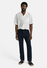 Korte mouwen, lichtgekleurde shirt met een gestructureerd patroon, gecombineerd met donkere, loszittende broeken en eenvoudige sandalen.