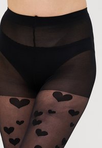 Svart genomskinlig strumpbyxa med hög midja, med ett mönster av svarta hjärtformer över hela ytan och en slät, mjuk textur.