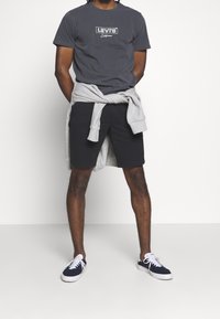 Šedé tričko Levi's s logem, šedá mikina uvázaná kolem pasu, černé šortky a námořnické tenisky s bílou podrážkou. Bílý pozadí.
