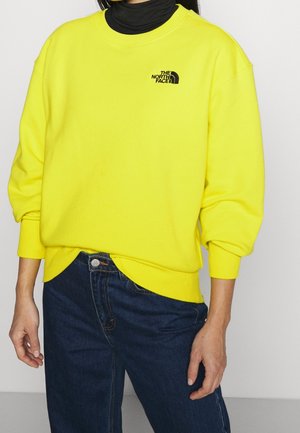 Osoba nosząca jaskrawożółtą bluzę The North Face na czarnym golfie, w połączeniu z ciemnoniebieskimi dżinsami, stojąca na jednolitym tle.