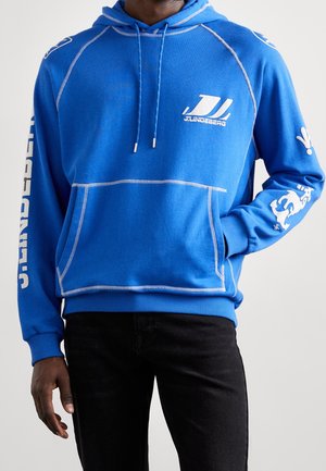 Mann trägt blaues J.Lindeberg Hoodie mit weißem Logo und Text auf Brust und Ärmeln, kombiniert mit schwarzen Hosen, Hand in der Tasche.
