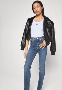Veste bomber cropped en cuir noir sur un débardeur blanc avec un logo ; associée à un jean skinny bleu taille haute. Textures lisses et design ajusté.