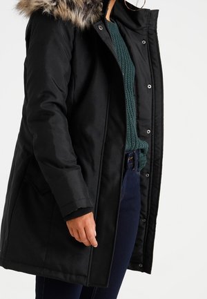 Parka nera con cappuccio foderato in pelliccia, chiusure a bottone e a pressione, tasche laterali e design aderente. Indossata sopra un maglione verde lavorato a maglia.