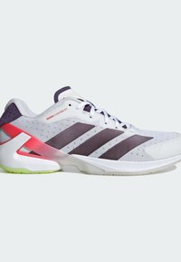 Baskets blanches avec des rayures noires Adidas, un support de talon rouge, des lacets blancs et une semelle vert clair sur fond uni clair.