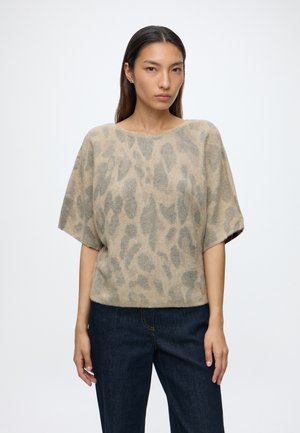 Frau trägt einen beige-grauen kurzärmeligen Pullover mit Leopardenmuster und dunkelblaue Jeans, steht vor einem schlichten weißen Hintergrund.