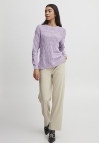 Maglione a maglia viola chiaro con un motivo testurizzato, abbinato a pantaloni beige a gamba larga e stivaletti neri alla caviglia con dettagli bianchi.