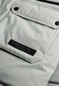 Jasnoszara marynarka sportowa o gładkiej teksturze. Posiada kieszeń z klapą, czarną metkę z logo oraz czarne akcenty z metalowymi elementami.