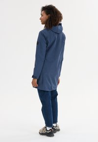 Pitkä tummansininen takki, jossa on huppu, napitettavat hihansuut ja teksturoitu kangas. Yhdistetty sinisiin farkkuihin ja harmaisiin lenkkareihin.