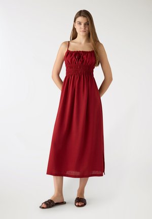 Giovane donna in piedi con le mani dietro la schiena, indossa un vestito midi rosso senza maniche con corpino arricciato e sandali neri bassi su sfondo bianco.