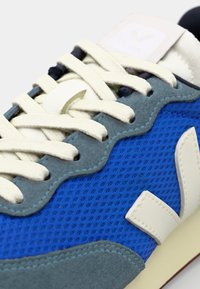 Blå mesh-sneaker med grå semsket skinn-detaljer, kremfargede lisser og en hvit logo-patch på tungen. Strukturert stoff og synlige sømmer.