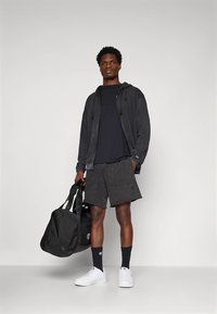 Must kapuutsiga pusa, tume t-särk ja lühikesed püksid, kõik tekstuurse materjaliga. Valged tossud, mustad sokid ja must duffelkott täiustavad välimust.